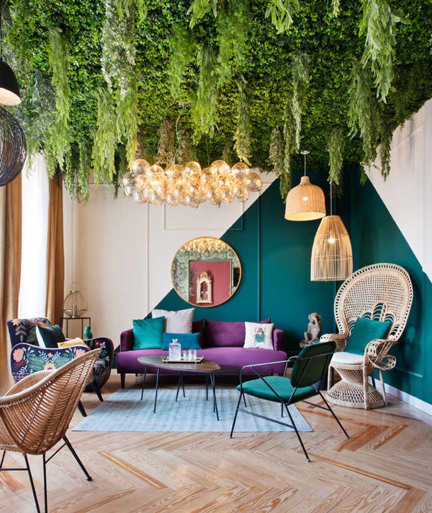 Living con muebles enterciopelo y mimbre, en colores lila, verde y azules. Lámparas de mimbre y vidrio y cielo cubierto de plantas y hojas verdes.