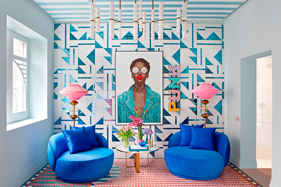 Sala de estar con muro con diseños geométricos en vidrio, un gran cuadro de una modelo de color , dos sillones individuales de terciopelo azul, cada uno con una lámpara de pie en tonos rosados y cobre.