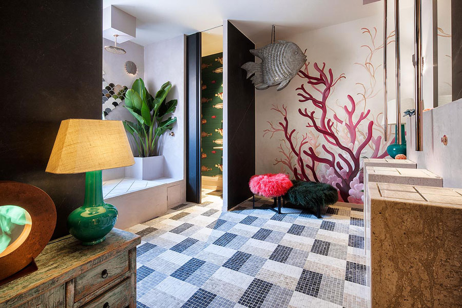Vista de baño con piso de mosaico, y decoración marina, con un muro pintado con diseño de coral, plantas tropicales y una figura de un pez en tonos grises.
