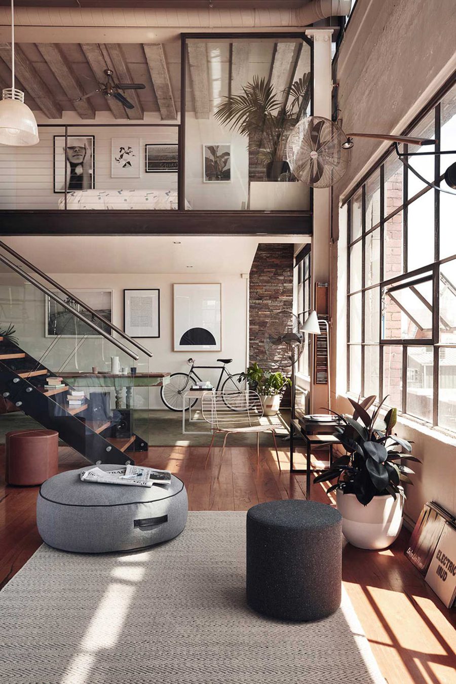 Casa de estilo industrial con espacios abiertos y grandes ventanales. Hacia el fondo hay una mesa de comedor, una bicicleta, tres cuadros en blanco y negro, y un muro de ladrillo. Adelante hay tres pouf de tonos neutros, una planta con macetero blanco y una alfombra blanco crudo.