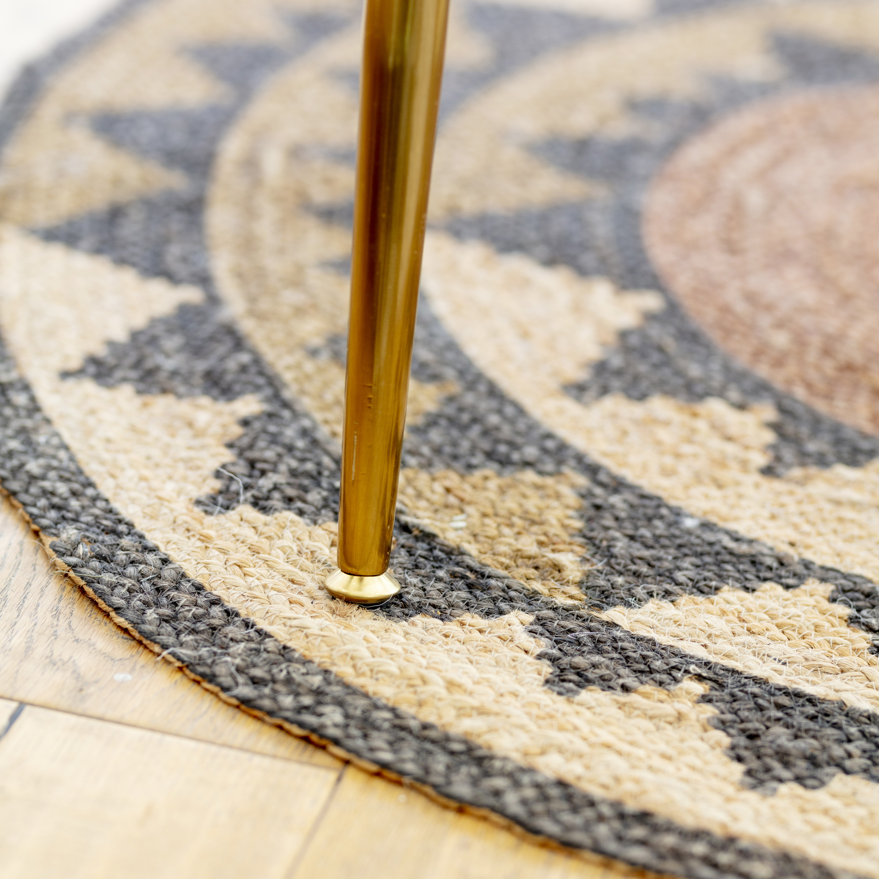 Pata cónica dorada de una mesa, apoyada sobre una alfombra de yute con estampado negro y tonos cálidos. El piso es de madera clara.