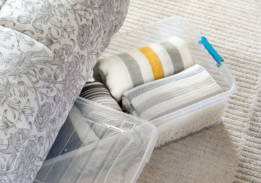 Caja organizadora de plástico transparente con mantas en su interior. Se está asomando desde la parte de abajo de una cama.