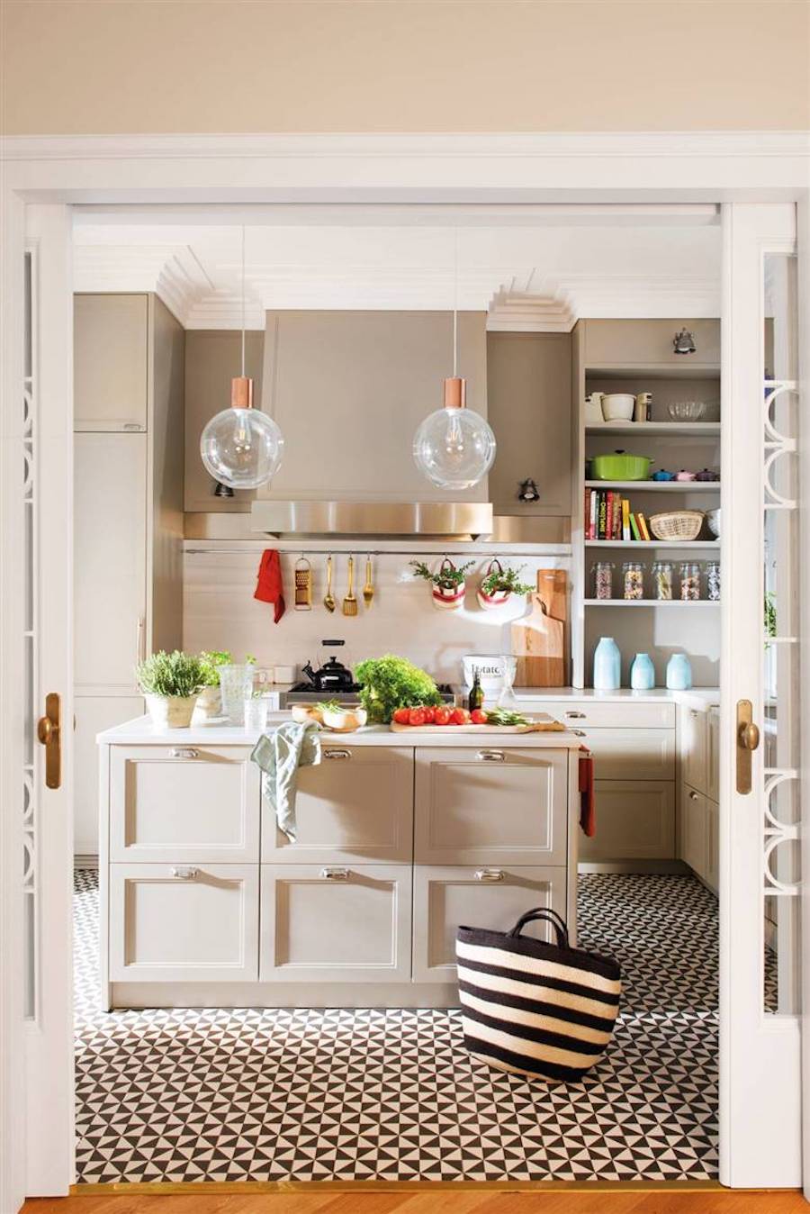 Cocina con isla y piso vinílico con patrón geométrico en blanco y negro. Sobre la isla hay diferentes verduras y utensilios de cocina. Desde el techo cuelgan dos lámparas con forma de esfera transparentes.