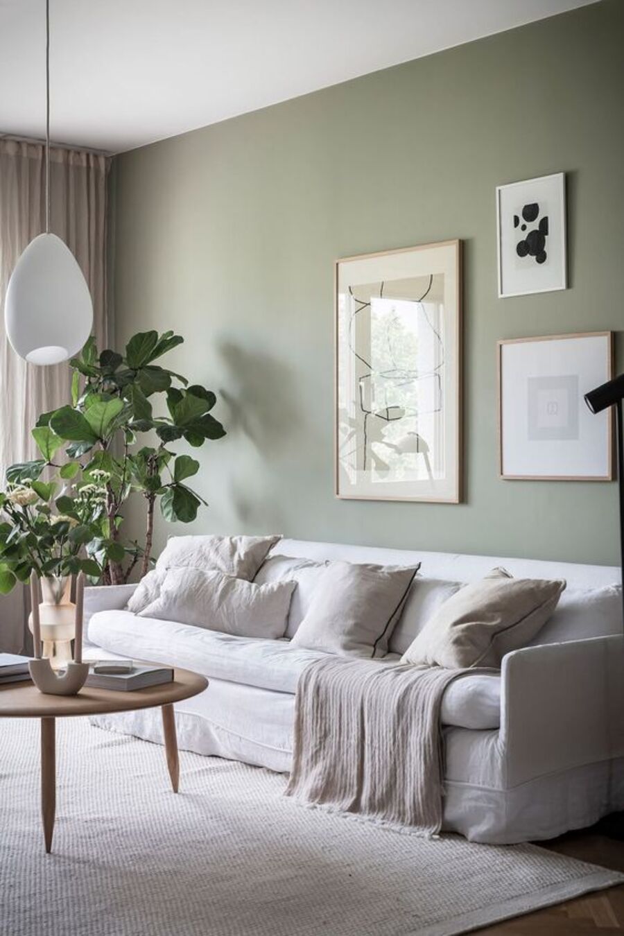 Sala de estar con alfombra, sofá y cojines blancos, cortinas beige y muro verde claro. En este último hay tres cuadros con ilustraciones minimalistas. Frente al sofá hay una mesa de centro circular de madera con adornos blancos y planta en su superficie.