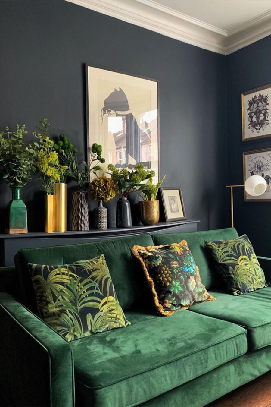 Sala de estar de paredes negras y sofá verde esmeralda con cojines con hojas tropicales verdes. Detrás del sofá hay un mueble negro que sostiene seis floreros de distintos colores y formas, los cuales contienen hojas verdes de diferentes tipos. 