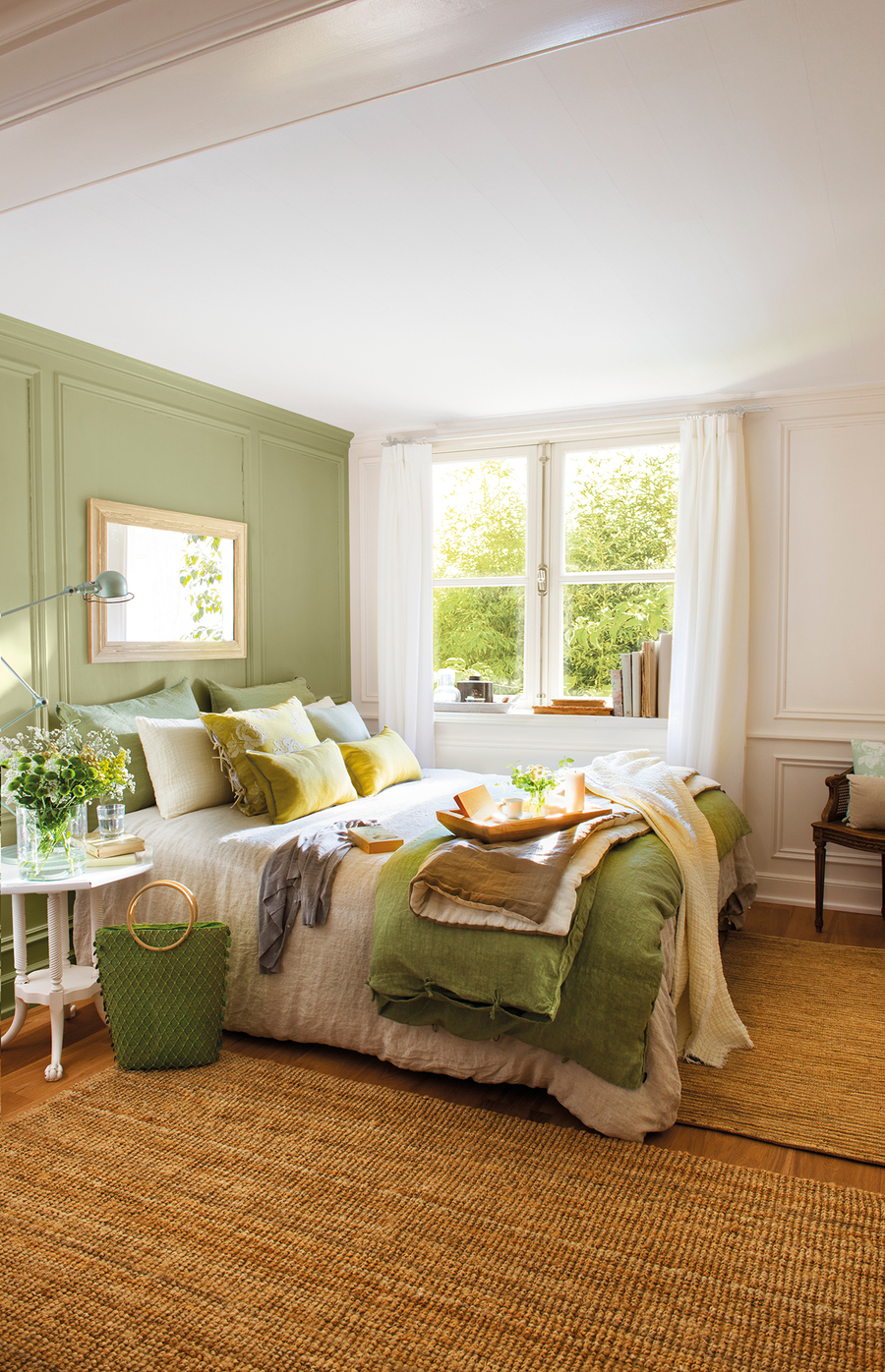 Dormitorio de techo blanco, piso de madera, pared blanca y pared de acento verde claro. La ropa de cama es de diferentes tipos de verde, incluyendo los siete cojines y mantas sobre ella. Sobre el suelo hay dos grandes alfombras de yute.