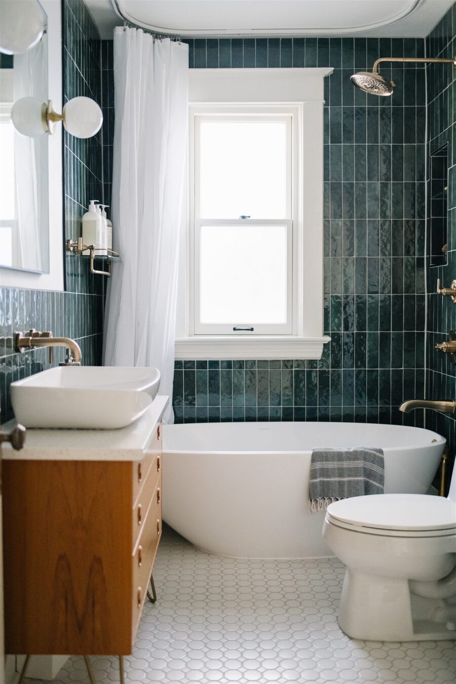 Baño con tina blanca con forma ovalada, azulejos verde petróleo y mueble del lavamanos de madera. El lavatorio, el piso y la cortina de la ducha son blancas, mientras que la grifería es dorada.