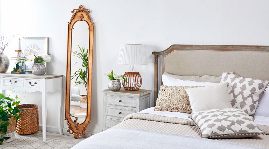 Dormitorio de estilo clásico con una cama con respaldo de madera y tapiz beige, cojines en tonos crema y ropa de cama beige y blanca. A su lado hay un velador y junto a éste, un espejo de cuerpo completo con marco en color cobre y diseño clásico. 