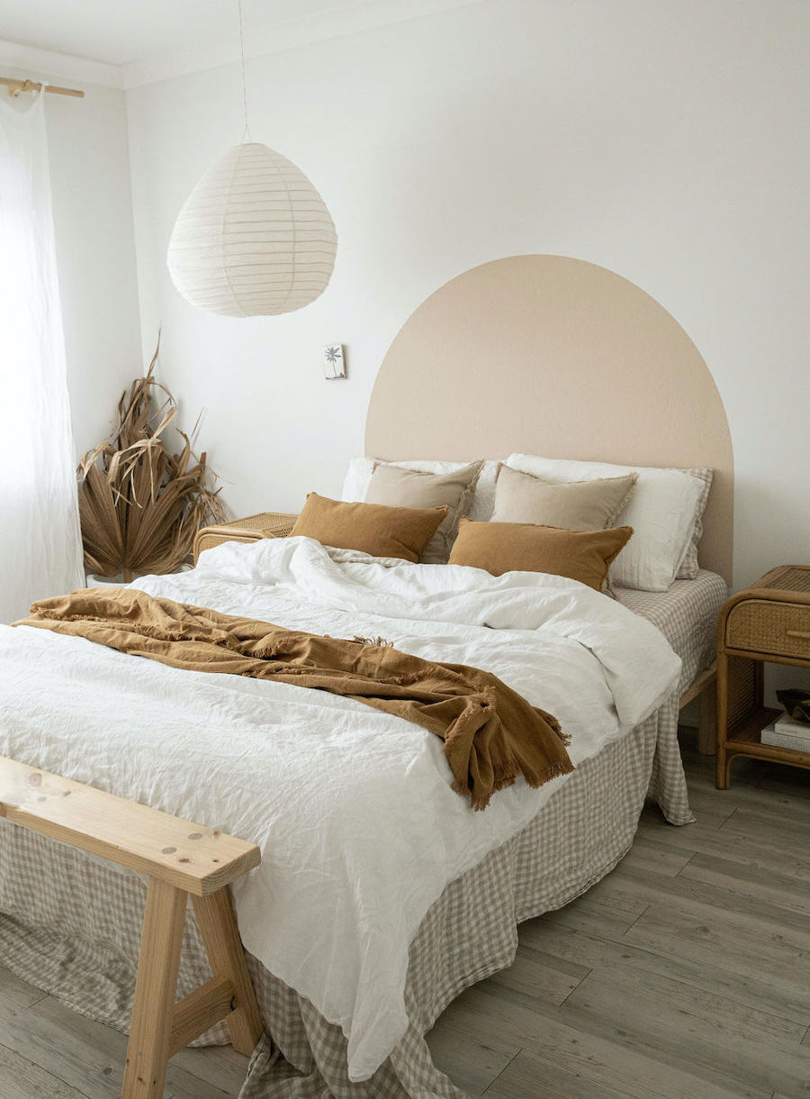 Dormitorio de muros blancos con un arco pintado en color beige, el cual juega como respaldo de cama. Los cojines son de tonos tierra, el plumón de la cama es blanco y hay una manta café sobre él.