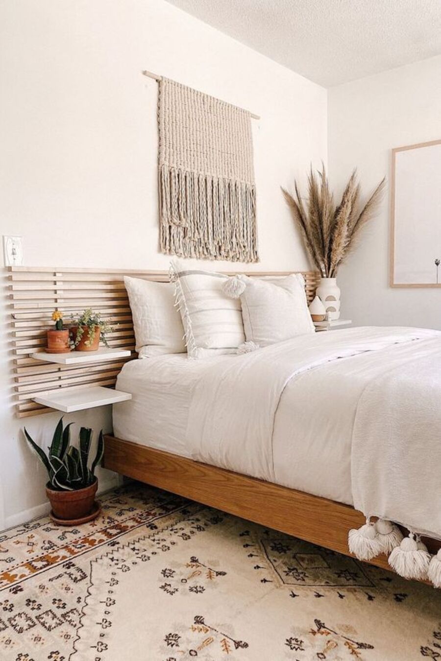 Dormitorio con paredes blancas y alfombra con diseño en tonos tierra. La ropa de cama también es blanca, el respaldo y la base son de madera y hay algunas plantas en los veladores y una en el suelo. 