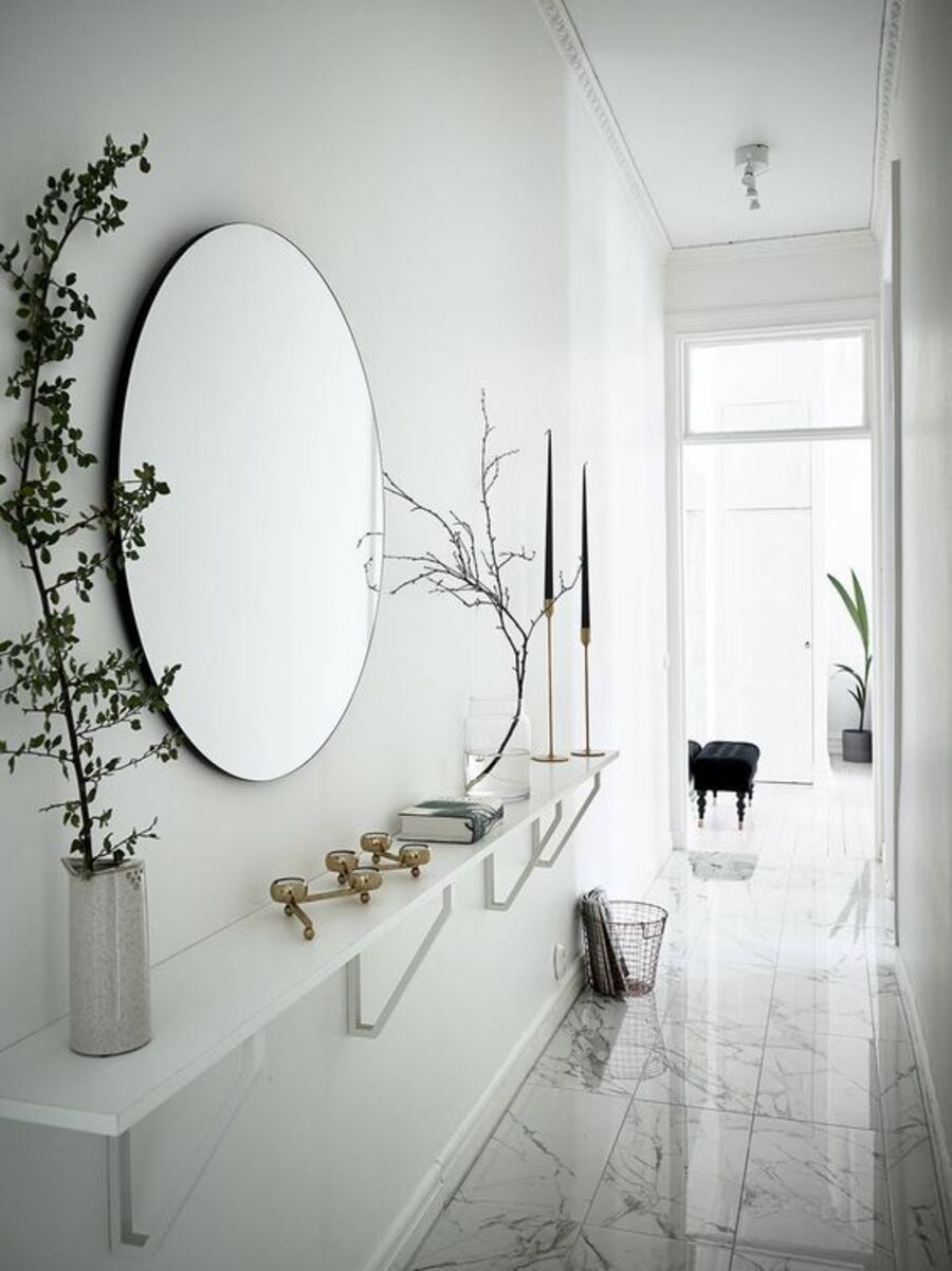 Pasillo blanco con un gran espejo redondo en uno de sus muros. Debajo hay una repisa larga y blanca que sostiene diversos adornos dorados y dos floreros con ramas.