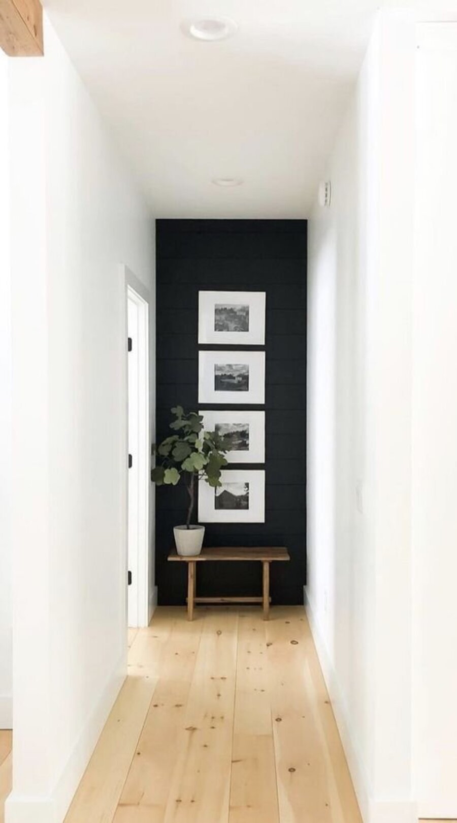 Largo pasillo de paredes blancas y piso de madera clara. La pared del fondo está pintada negra y de ella cuelgan cuatro fotografías con marco negro. En el suelo hay una pequeña banca de madera que sostiene una planta.