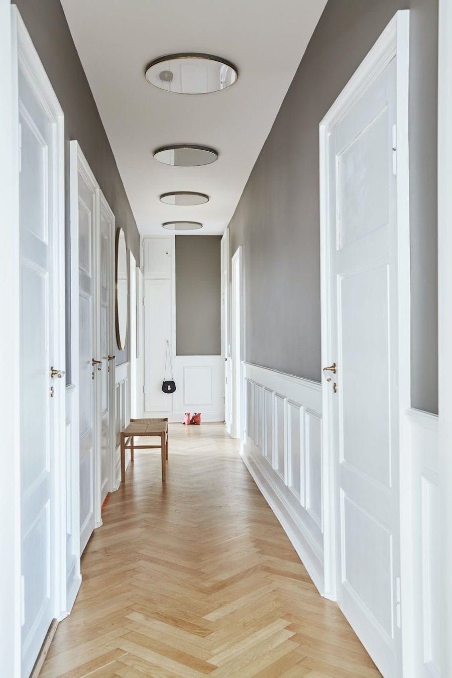 Largo pasillo de piso de madera, techo blanco con plafones tipo espejo y paredes grises con molduras blancas. Las puertas también son blancas.