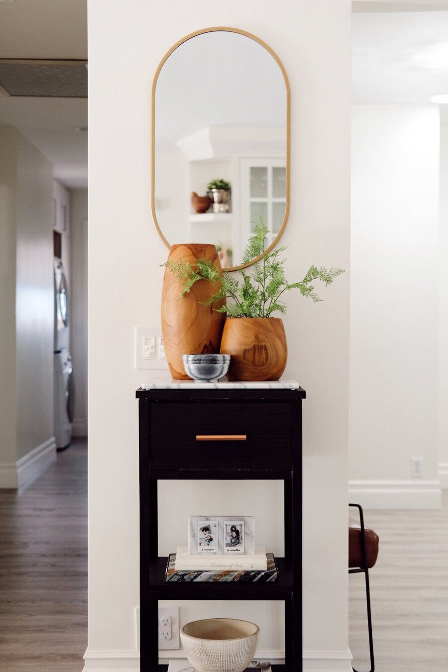 Pequeño recibidor con un mueble de apoyo negro con un cajón con tirador en color cobre. Sobre él hay un dos floreros de madera, el más pequeño de ellos sostiene una planta. De la pared cuelga un espejo ovalado con marco dorado.
