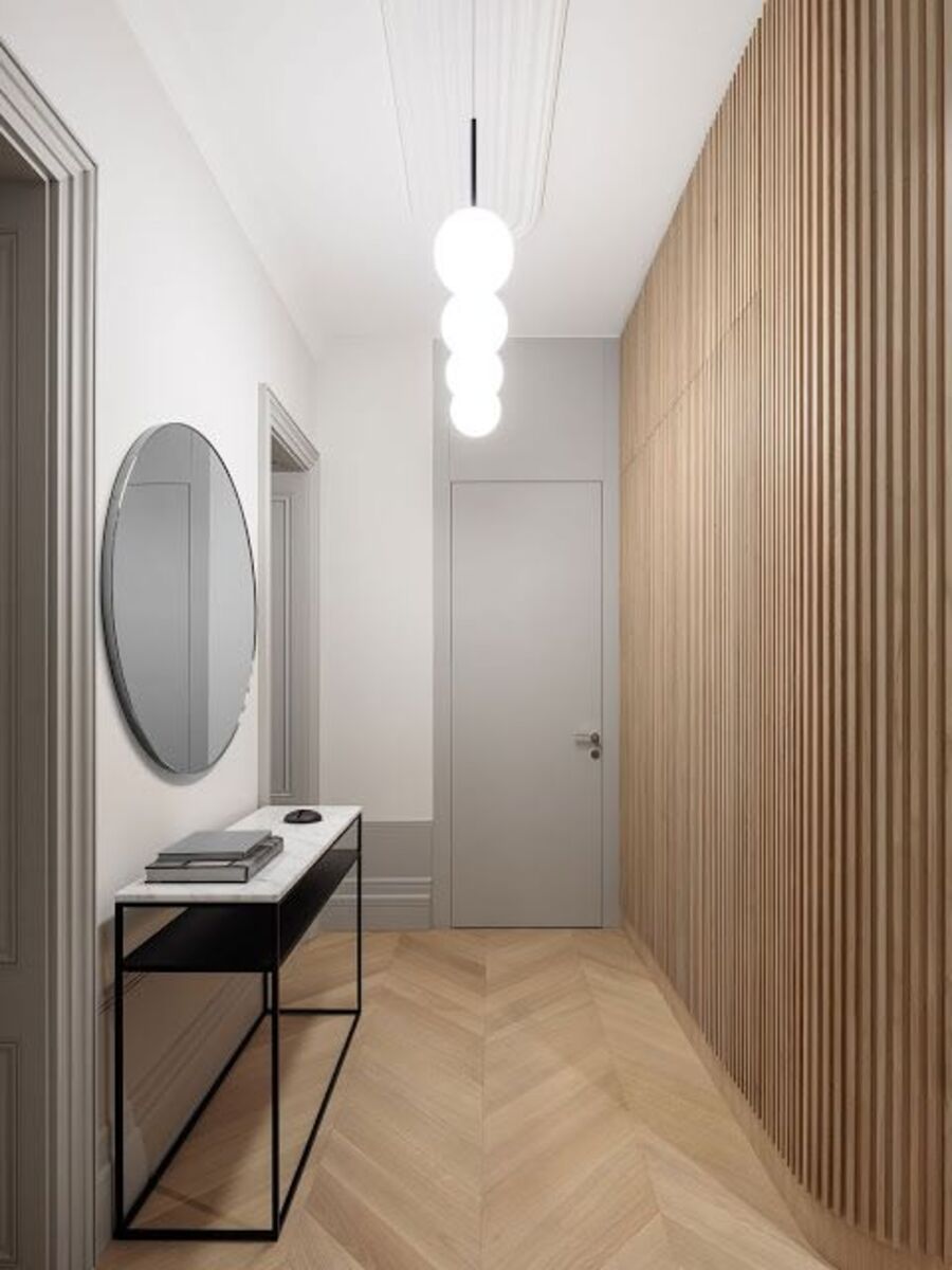 Pasillo de piso de madera y techo blanco. Uno de los muros está cubierto por un revestimiento con listones de madera, mientras que la otra pared es blanca con molduras grises. Tiene dos puertas y entre ellas, un gran espejo redondo con un arrimo negro.