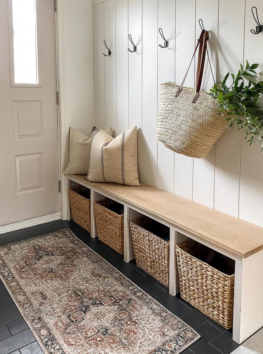 Recibidor con un mueble de madera estilo banqueta con espacio para poner canastos de fibras naturales debajo. Sobre él hay dos cojines en un tono crudo. En la pared hay cinco percheros, uno de ellos sostiene un bolso con un planta que sale de su interior. El piso es negro y sobre él hay un alfombra estampada en tonos rojizos.