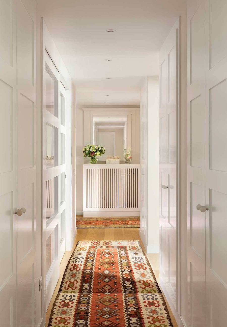 Pasillo de muros y puertas blancas y una larga alfombra con un estampado en colores rojizos. 