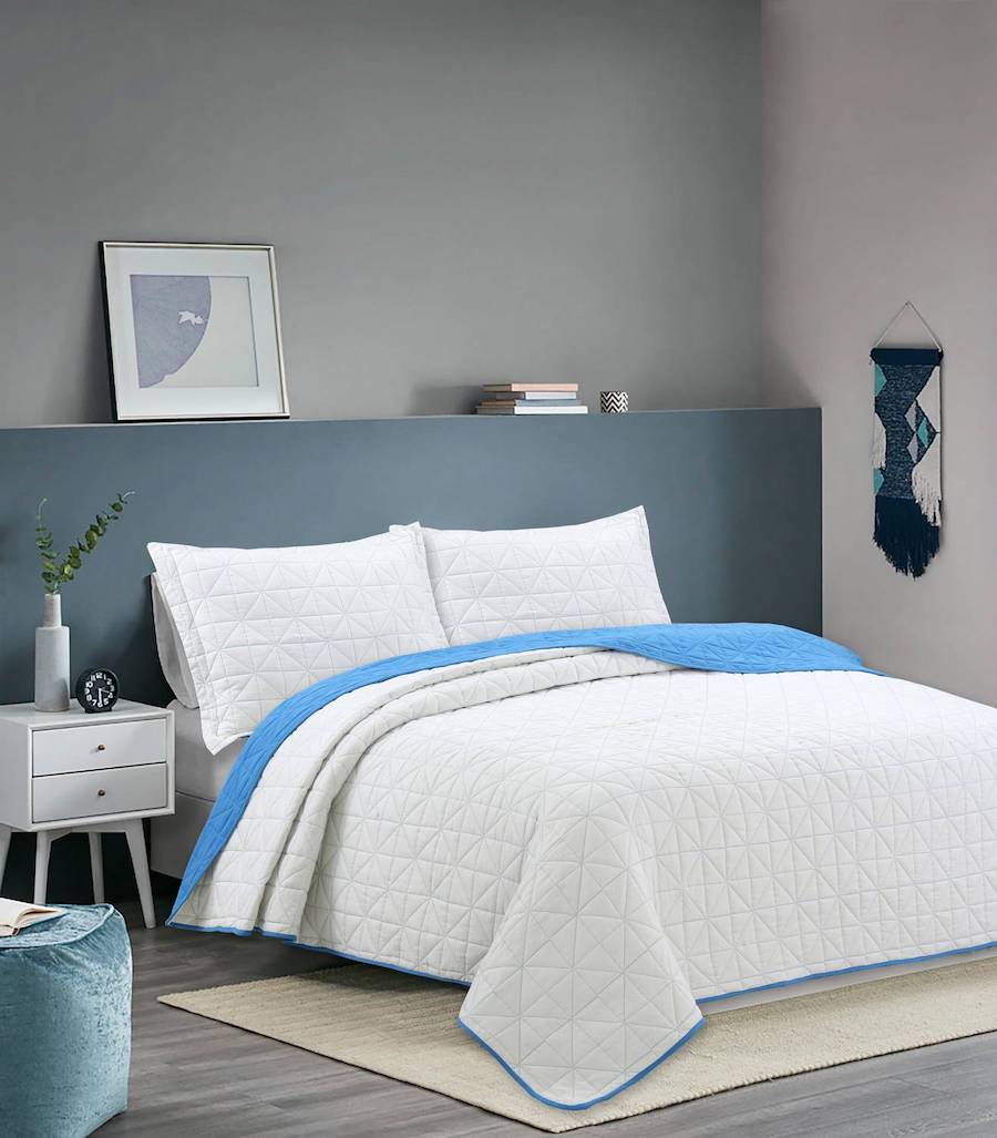 Dormitorio en tonos azulados con un quilt blanco con detalles azules y almohadas blancas. Como respaldo hay una parte del muro que sobre sale y está pintada de un color azul petróleo. Sobre él hay un cuadro y una pila de libros. El resto del muro es gris.