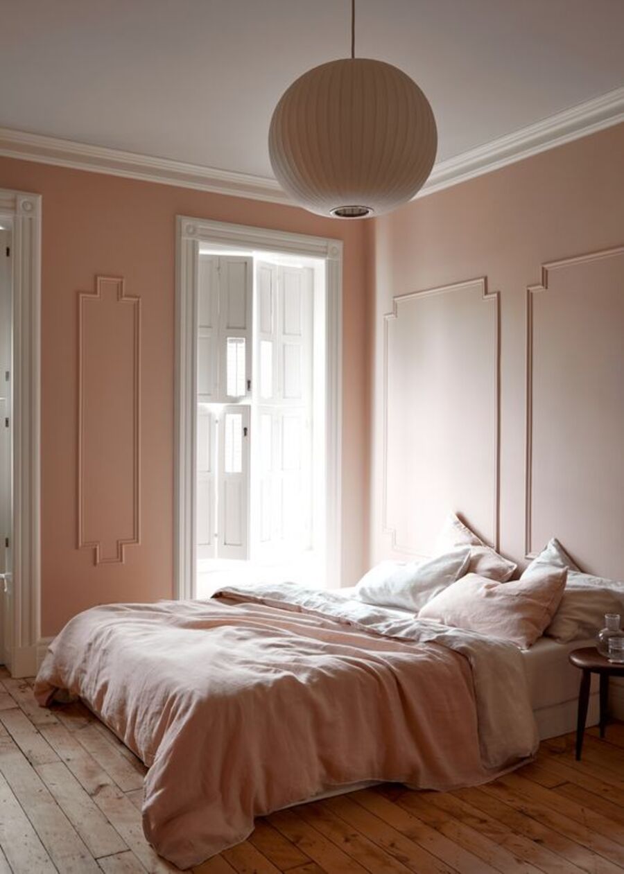 Habitación de muros rosa palo con molduras. El techo es blanco y el piso de madera. Desde el techo cuelga una gran lámpara redonda blanca. Debajo está la cama, cuyo plumón es rosado, así como también algunos cojines. Los otros con blancos, al igual que las sábanas.