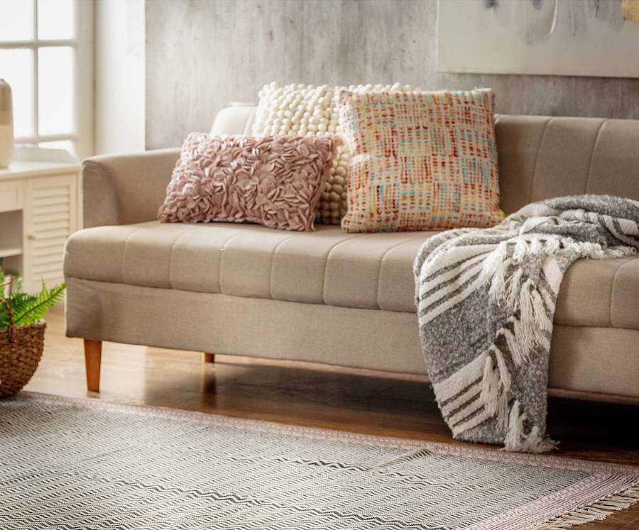 Detalle de un living con un sofá beige con cojines en tonos pasteles y una manta rayada gris con blanco. En el piso hay una alfombra gris con blanco y detalles rosados en el borde. 