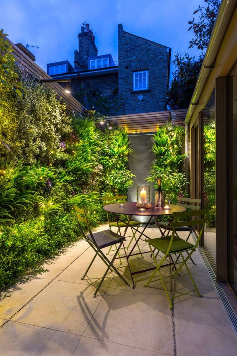 Terraza de una casa con jardines verticales y algunas luces que iluminan el atardecer. A un costado de las plantas hay una mesa de comedor con cuatro sillas metálicas verdes.