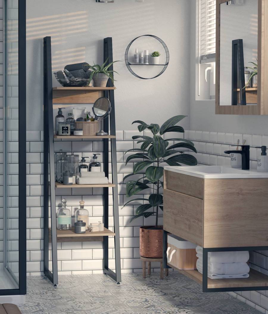 Baño de estilo moderno con paredes gris claro con azuejos blancos en la parte inferior. Hay una repisa tipo escalera con estructura negra y superficies de madera clara. Sobre ellas hay artículos de belleza e higiene, un espejo, una planta y toallas. A su lado hay una planta con macetero de cobre. En el muro del lado está el mueble de vanitorio y un espejo, ambos de madera clara.