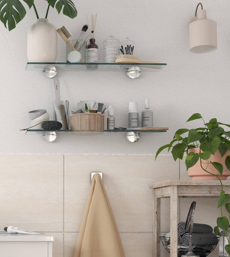 2 mini repisas flotantes de vidrio pegadas en el muro de un baño. Sobre ellas hay una planta, artículos de higiene y otros utensilios de belleza. Debajo hay una toalla beige colgando de un perchero, y a su lado, un pequeño estante con una planta.