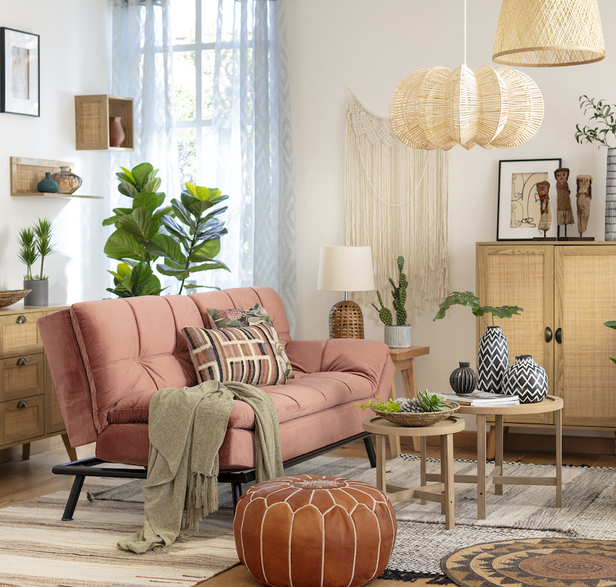 Sala de estar de estilo boho chic con un futón color salmón, alfombras en colores neutros, un pouf de cuero y muebles de madera clara con detalles en ratán. Del techo cuelgan 2 lámparas de fibras naturales.