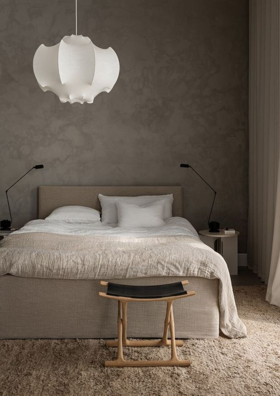 Dormitorio de paredes grises con textura, alfombra beige y una cama con sábanas y cojines blancos, y un cobertor beige. Sobre la cama cuelga una lámpara colgante blanca.