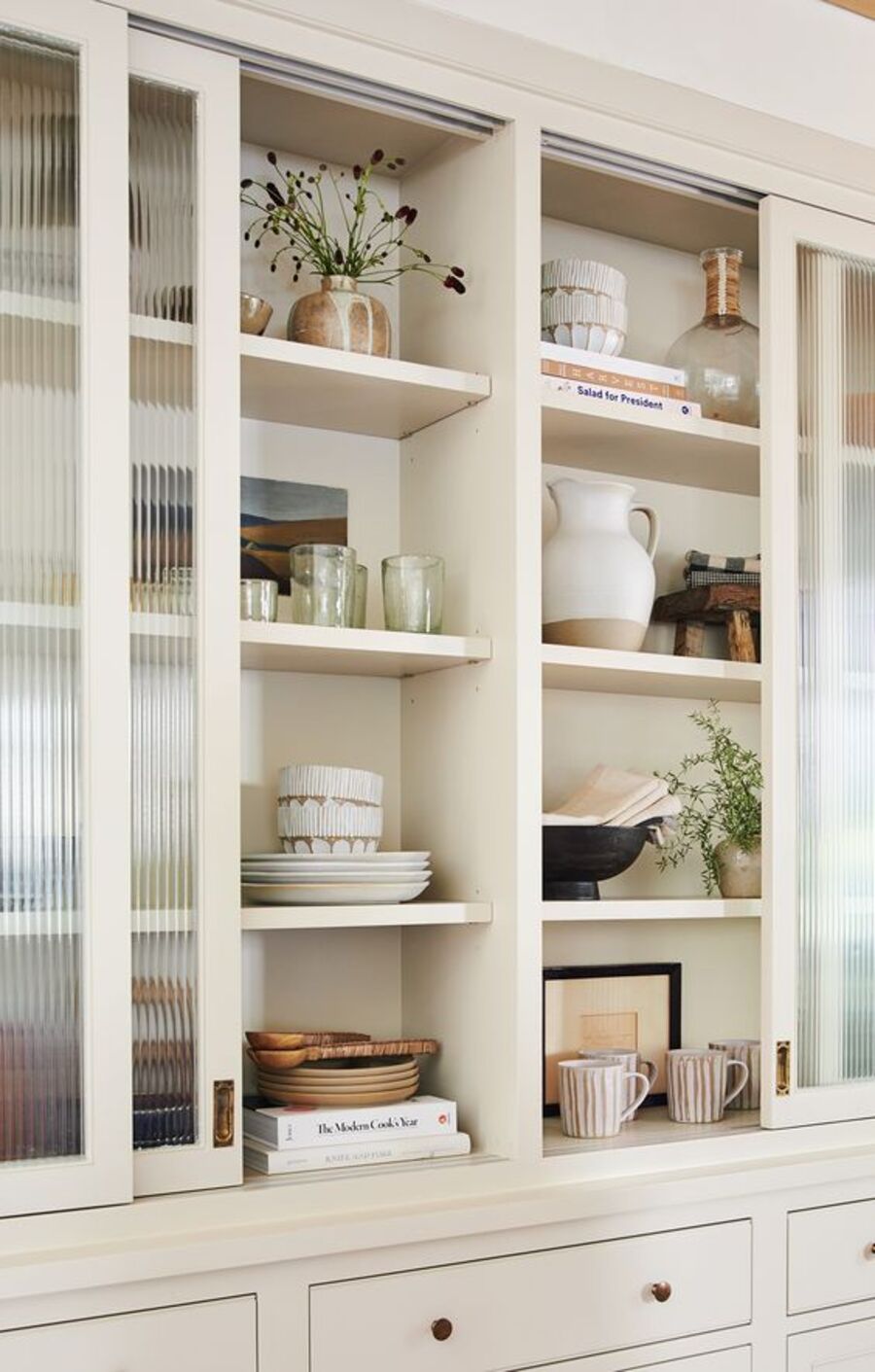 Mueble de comedor con varias repisas y puertas correderas de vidrio acanalado. En los estantes hay vasos, platos, tazas, bowls, jarrones, libros y plantas. En la parte inferior del mueble hay cajones.
