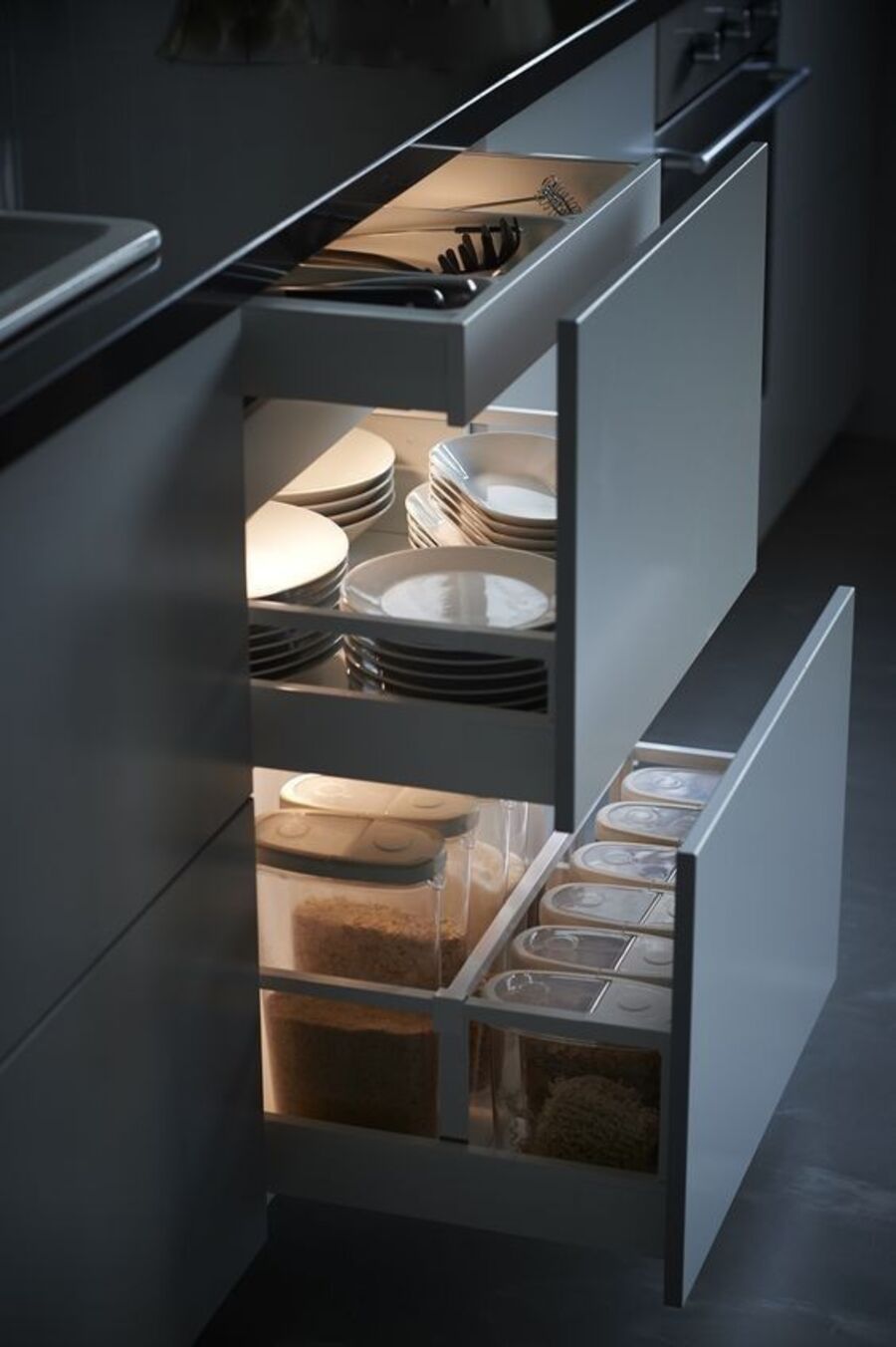 Detalle de un mueble de cocina gris con cubierta negra, cuyos cajones están semiabiertos y dejan ver diferentes contenedores, platos y utensilios. En cada cajón hay una luz que los ilumina.