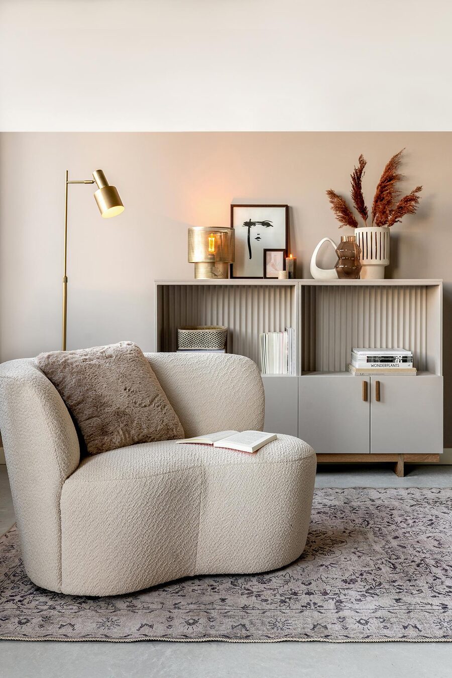 Sala de estar de tonos neutros con un sofá de un cuerpo de formas curvas y un mueble tipo biblioteca que sostiene libros, revistas, jarrones, floreros, cuadros y velas. A su costado hay una lámpara de pie dorada.